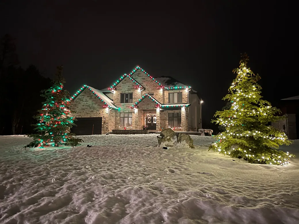 Stone home with red and green roofline lights and two lit evergreen trees in the snow